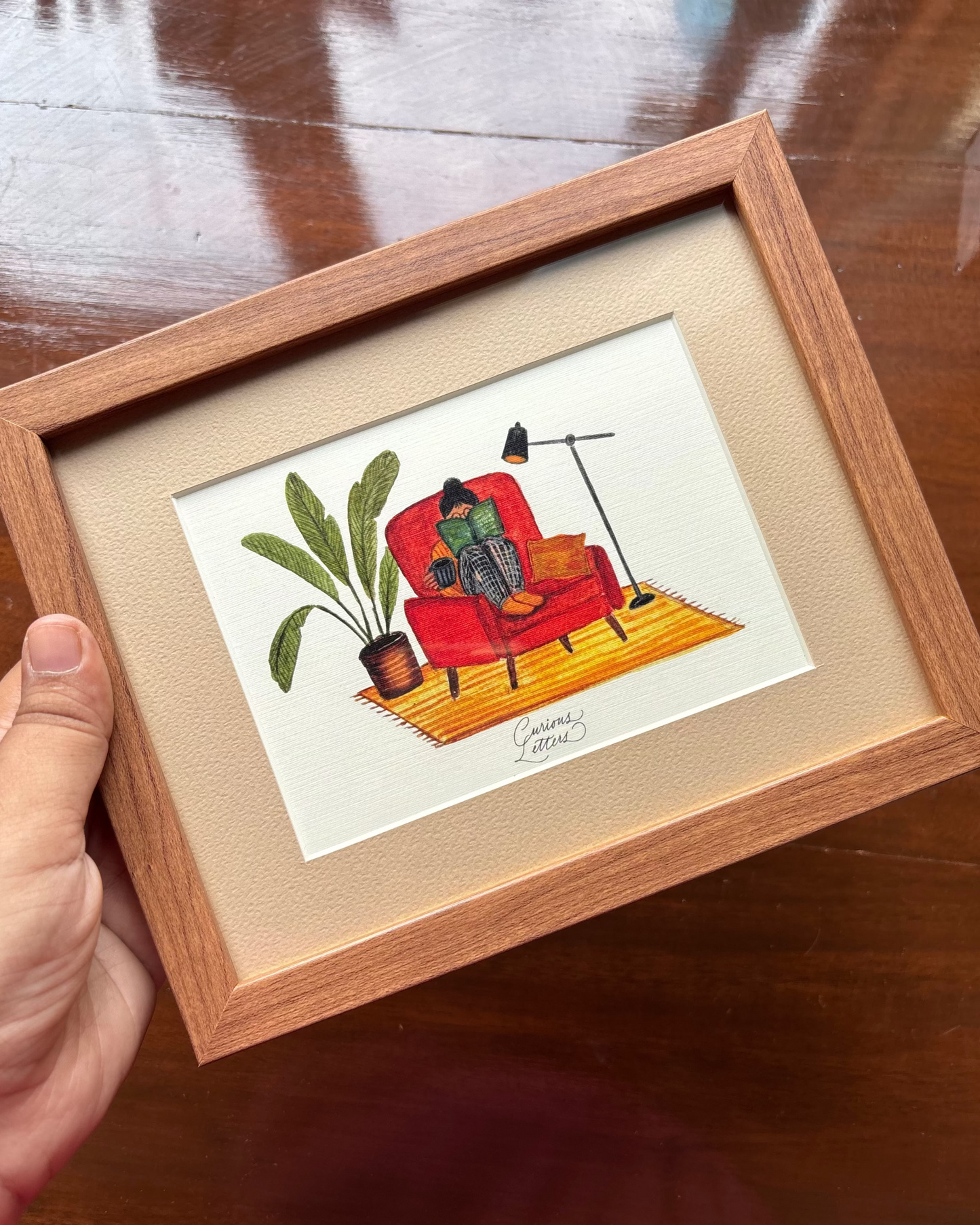 A framed art print featuring a hand-drawn illustration of a person sitting in a rust orange armchair reading a book, with a cup of tea in her hands, and a plant and floor lamp placed on the side. The image describes a cozy home feeling. Original art made by Curious Letters.