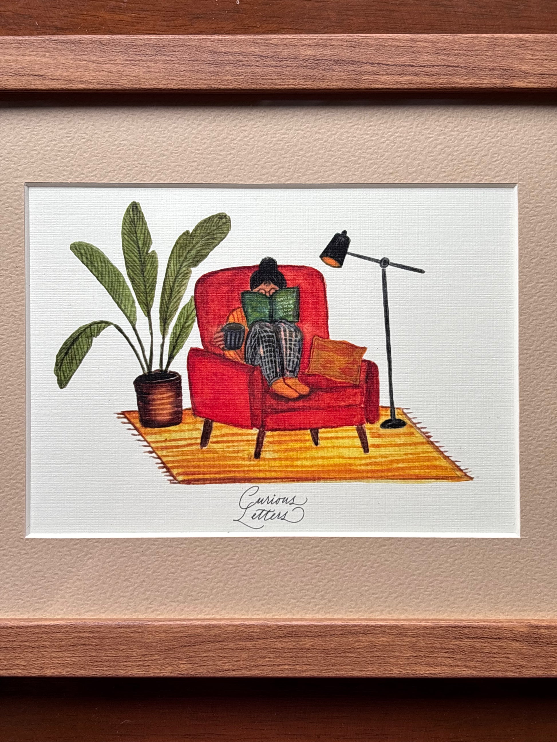 A framed watercolor painting of a cozy reading scene, featuring a person sitting in a red armchair reading a green book, next to a potted plant and a floor lamp, set within a white mat and a warm brown wooden frame. Original art made by Curious Letters.
