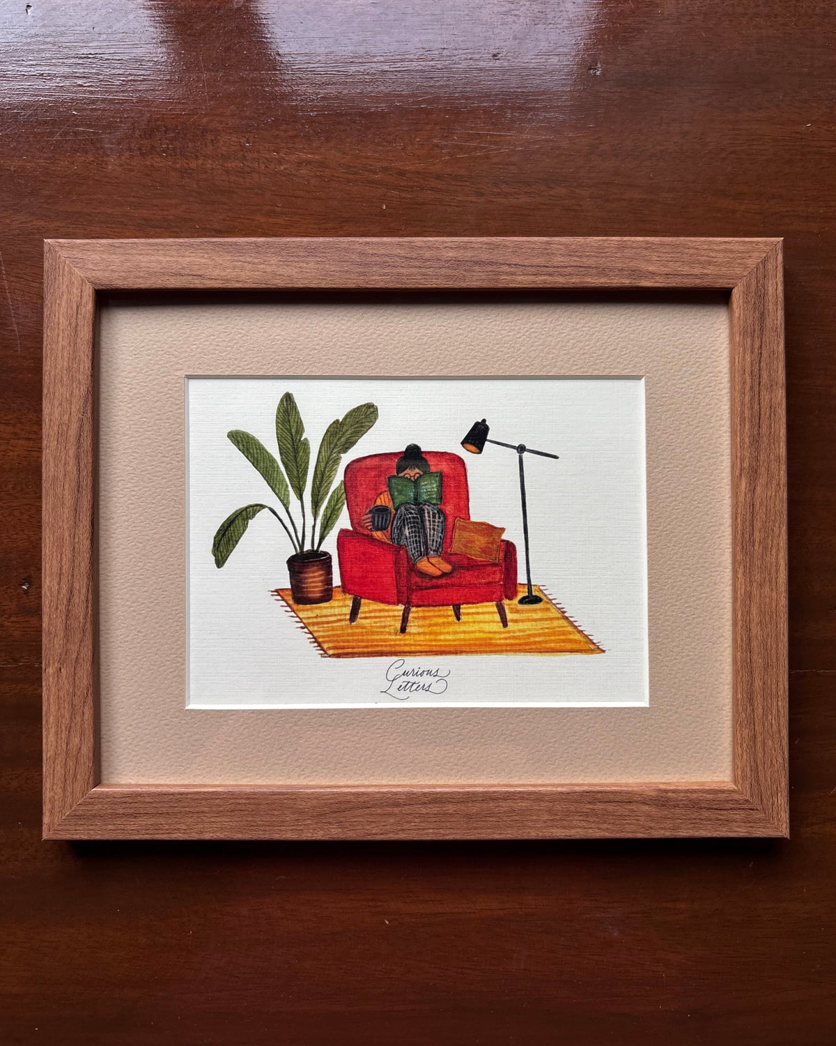 A framed art print featuring a hand-drawn illustration of a person sitting in a rust orange armchair reading a book, with a cup of tea in her hands, and a plant and floor lamp placed on the side. The image describes a cozy home feeling. Original art made by Curious Letters.