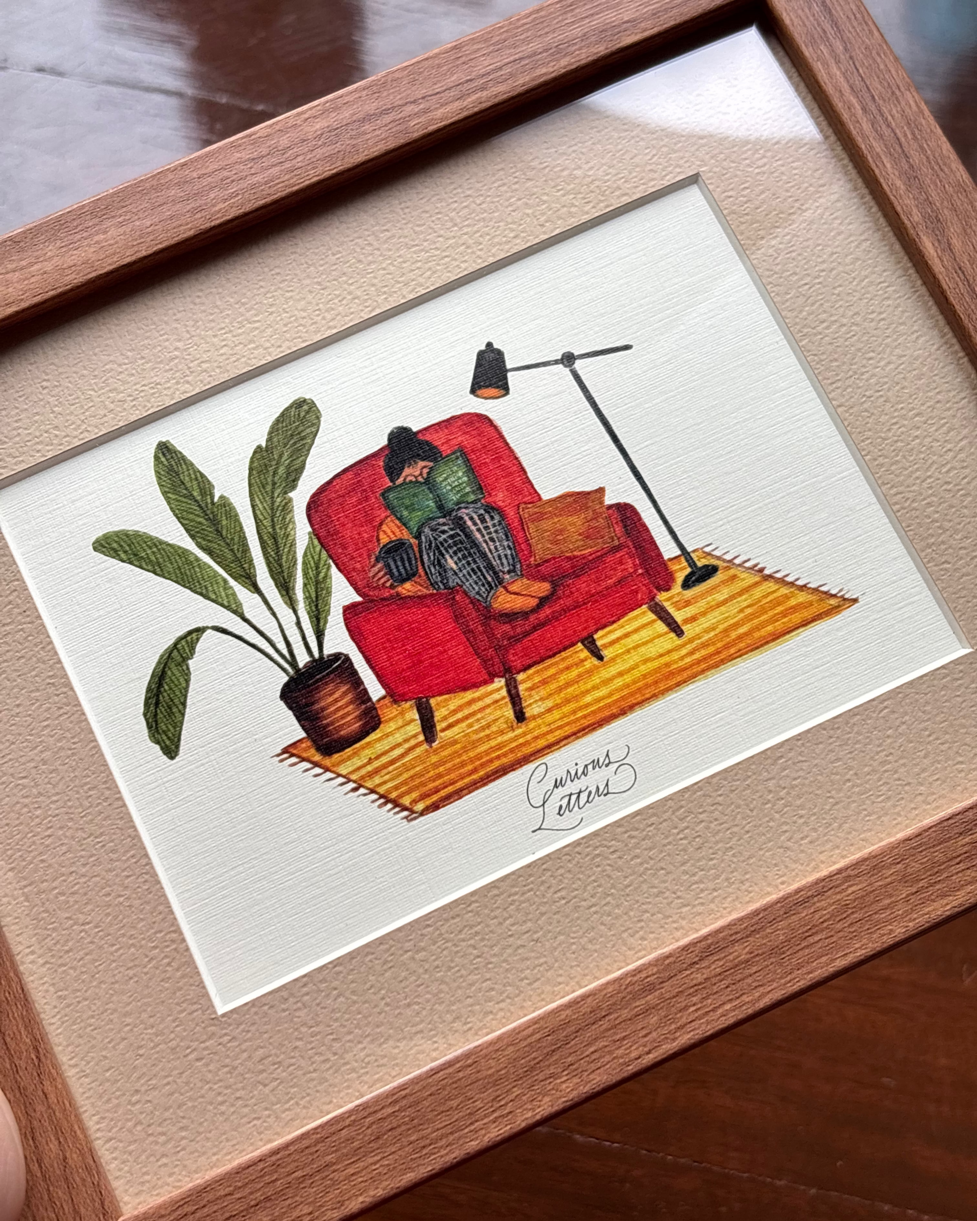 A framed art print featuring a hand-drawn illustration of a person sitting in a rust orange armchair reading a book, with a cup of tea in her hands, and a plant and floor lamp placed on the side. The image describes a cozy home feeling. Original art made by Curious Letters.