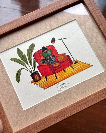 A framed art print featuring a hand-drawn illustration of a person sitting in a rust orange armchair reading a book, with a cup of tea in her hands, and a plant and floor lamp placed on the side. The image describes a cozy home feeling. Original art made by Curious Letters.