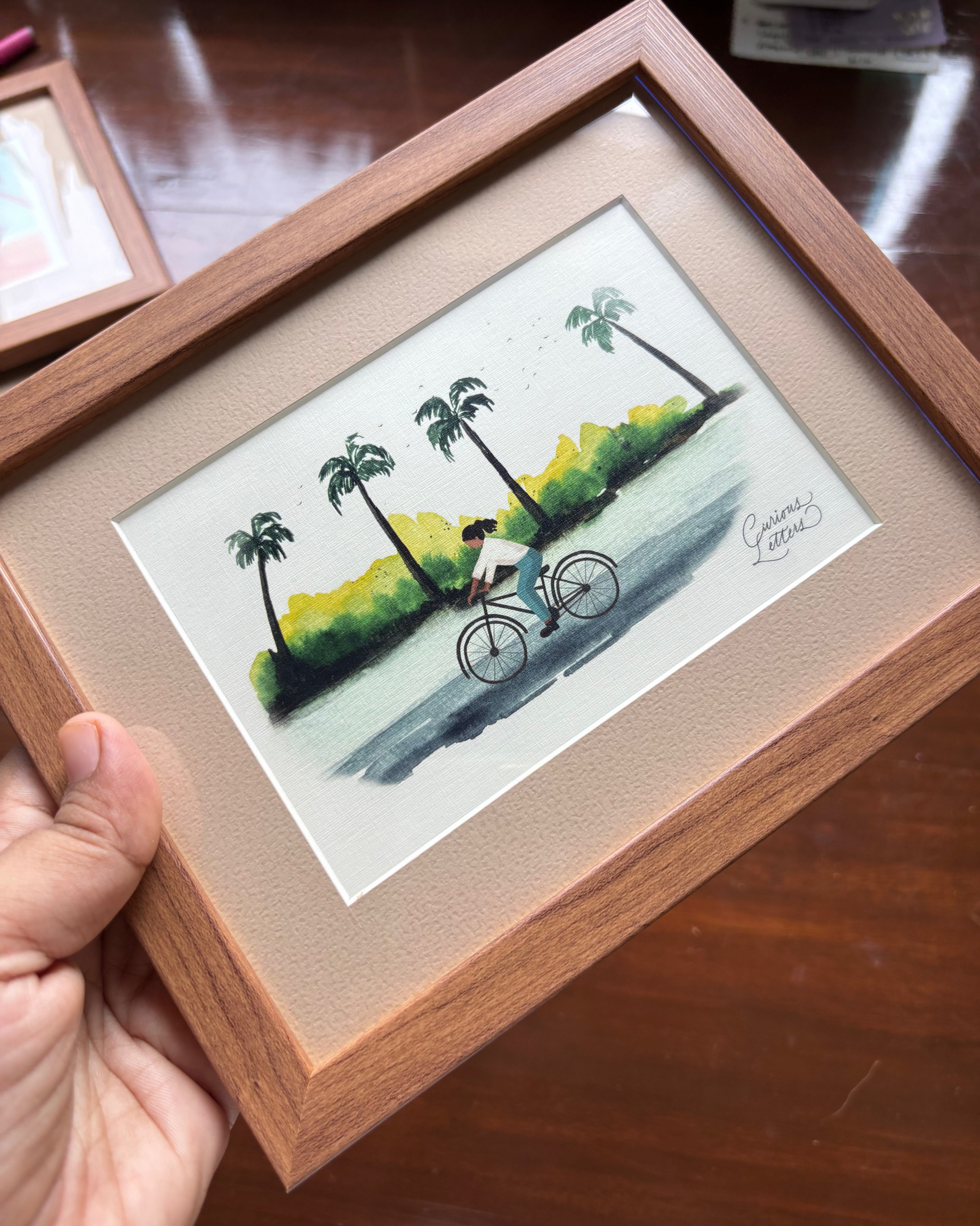 A framed watercolor painting of a girl riding a bicycle along a tropical shoreline with palm trees.