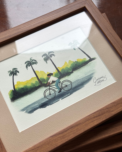 A framed watercolor painting of a cyclist riding along a path with palm trees, presented in a light brown wooden frame with a beige mat.