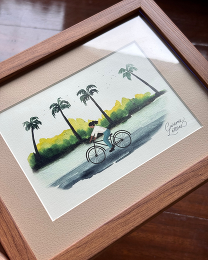A framed watercolor painting of a girl riding a bicycle along a tropical shoreline with palm trees.