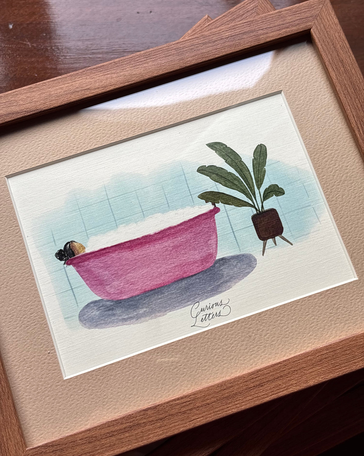 A framed watercolor painting of a girl in a pink bathtub full of soap bubbles, placed next to a potted plant, presented in a wooden frame.