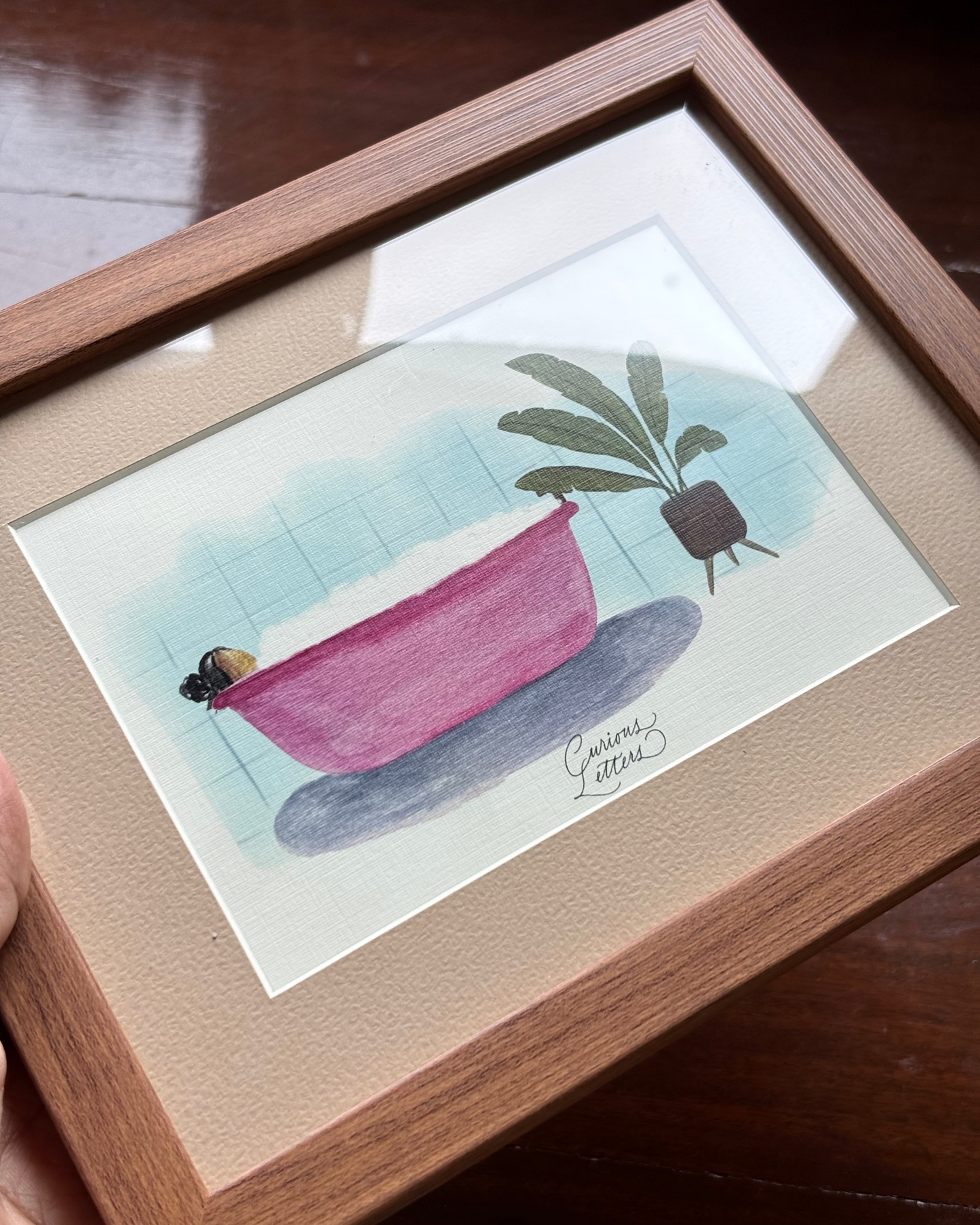 A framed watercolor painting of a girl in a pink bathtub full of soap bubbles, placed next to a potted plant, presented in a wooden frame.