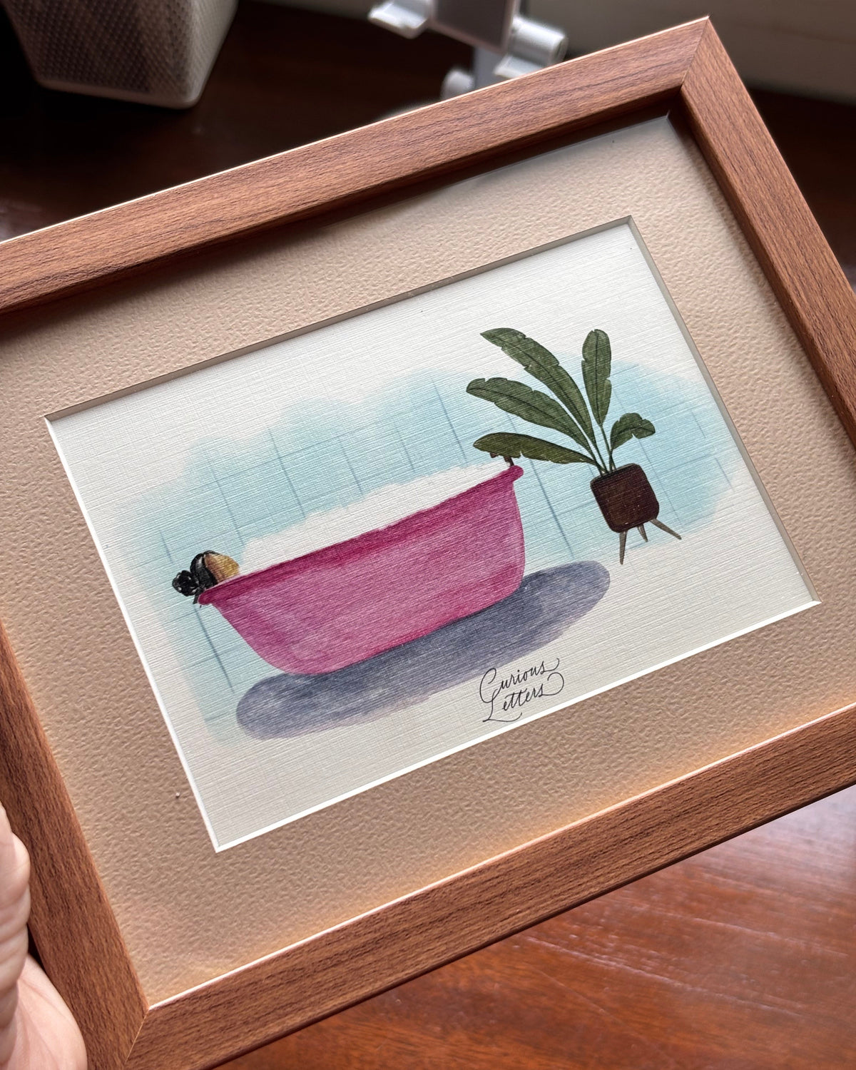 A framed watercolor painting of a girl in a pink bathtub full of soap bubbles, placed next to a potted plant, presented in a wooden frame.
