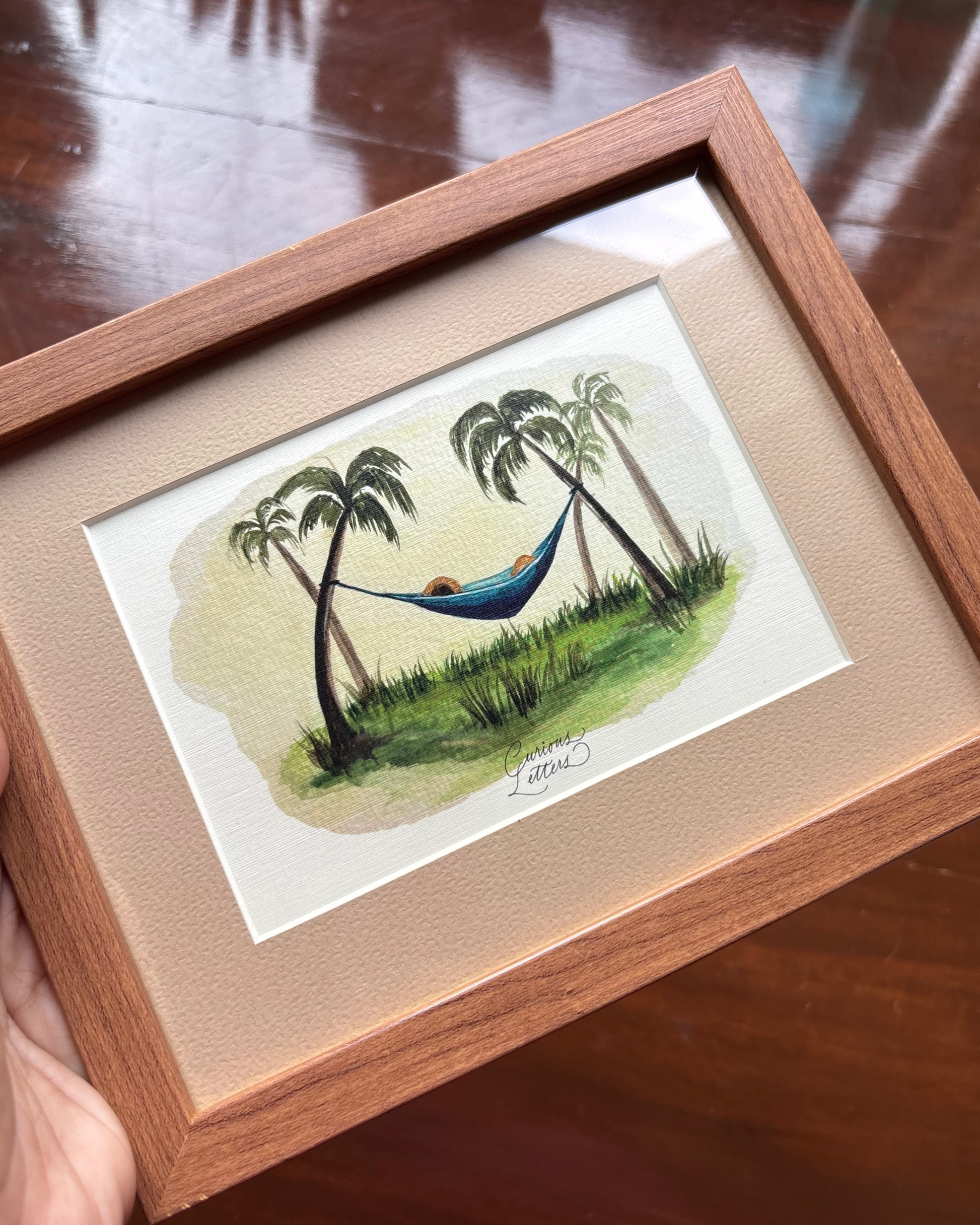 A framed watercolor painting of a tropical hammock scene with coconut trees, set in a light wood frame with a beige mat.