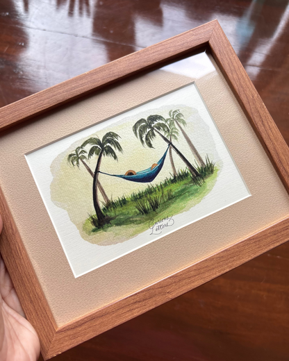 A framed watercolor painting of a tropical hammock scene with coconut trees, set in a light wood frame with a beige mat.