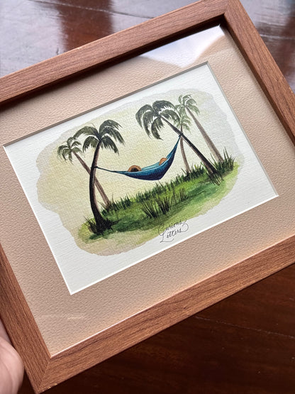 A framed watercolor painting of a tropical hammock scene with palm trees, green grass, and a blue hammock, presented in a light wood frame with a beige mat. 