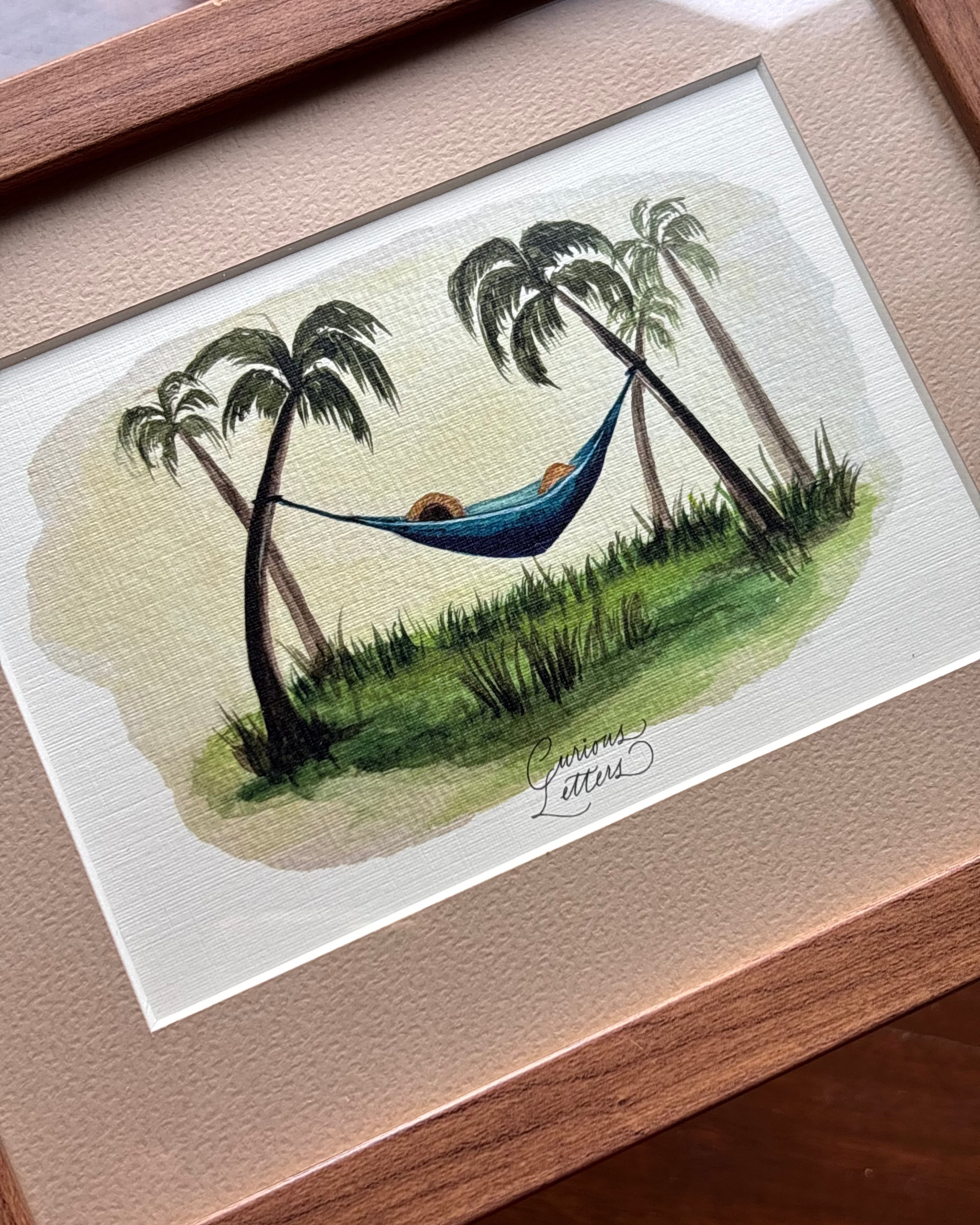 A framed watercolor painting of a relaxing tropical scene with palm trees, a blue hammock, and a person lying underneath, mounted on a textured beige mat inside a light wood frame.